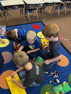 Group of five young boys sit together on a classroom carpet, building and playing with LEGO bricks.