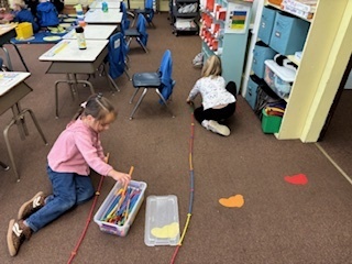 Two young girls in a classroom crouch on the floor, stretching long connecting sticks across the carpet while building.
