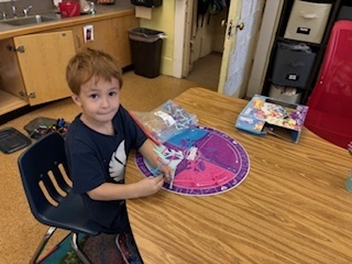Young boy with short brown hair sits at a round table, working on a colorful board game or activity mat with small pieces.