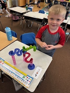 Young boy with blond hair stands at a desk, smiling proudly beside a colorful marble run toy he has built.