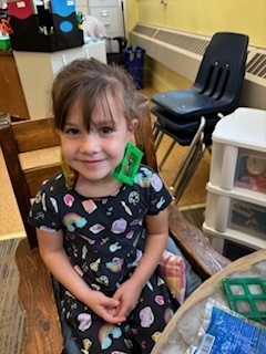 Young girl with brown hair in a ponytail smiles while sitting at a table, wearing a black dress with colorful designs. She playfully holds a green magnetic block to her ear like an earring.