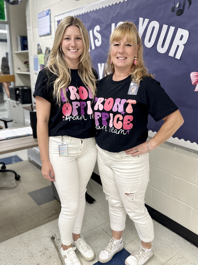 Two school staff members wearing matching black "Front Office Dream Team" shirts with white pants, smiling in a hallway.
