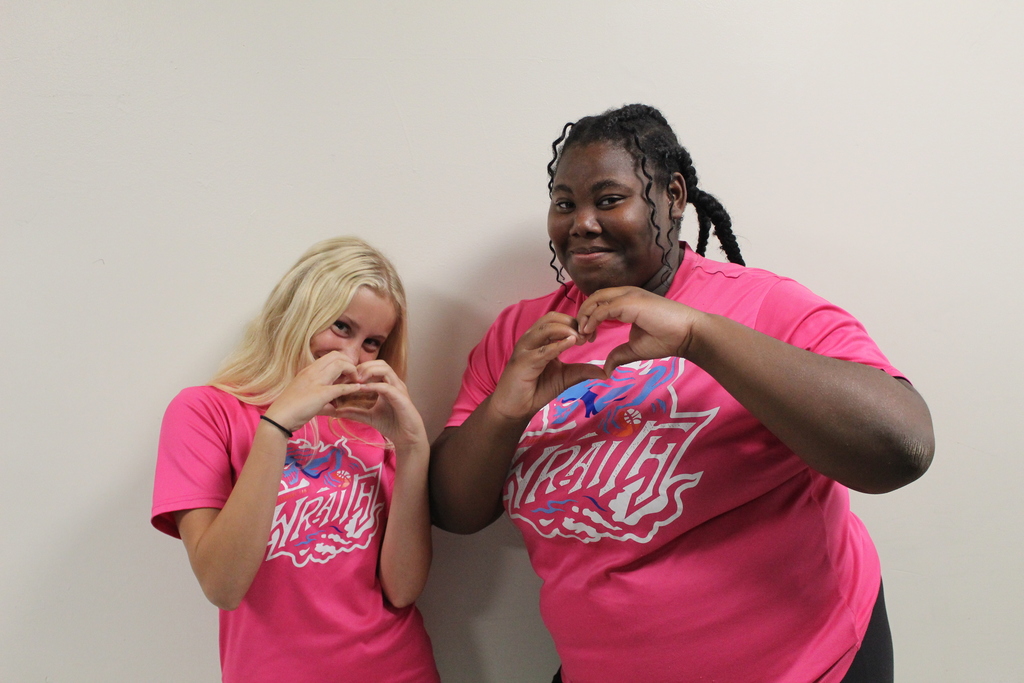 Two students in bright pink shirts forming heart shapes with their hands.
