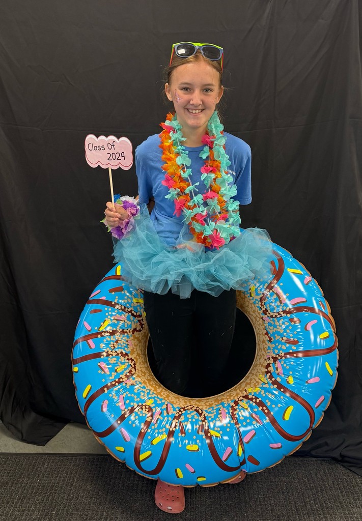 Student dressed for Beach Day. Wearing a blue shirt, tutu, colorful leis, and sunglasses while standing inside a large inflatable donut float and holding a “Class of 2029” sign.