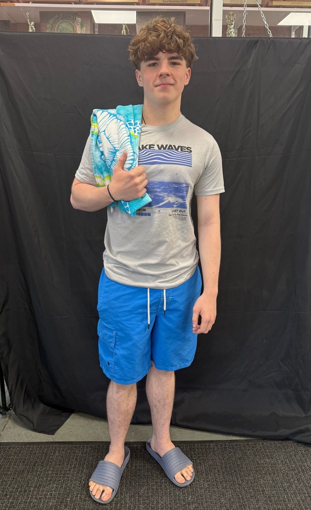 Student dressed for Beach Day. Wearing blue swim shorts, a gray “Lake Waves” t-shirt, and slide sandals, holding a folded beach towel over one shoulder.