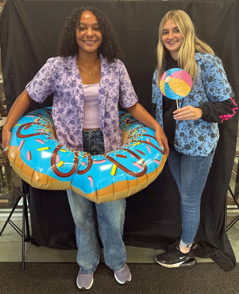 Two students dressed for Beach Day Spirit Week. One wears a purple floral shirt with jeans and stands inside a large inflatable donut float. The other wears a blue floral shirt and holds a paper beach ball prop.