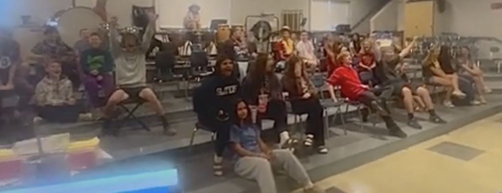 A large group of students sits on tiered risers in a music or band room. Many are smiling, some raising their arms in excitement. Musical instruments such as drums and percussion equipment are visible in the background.