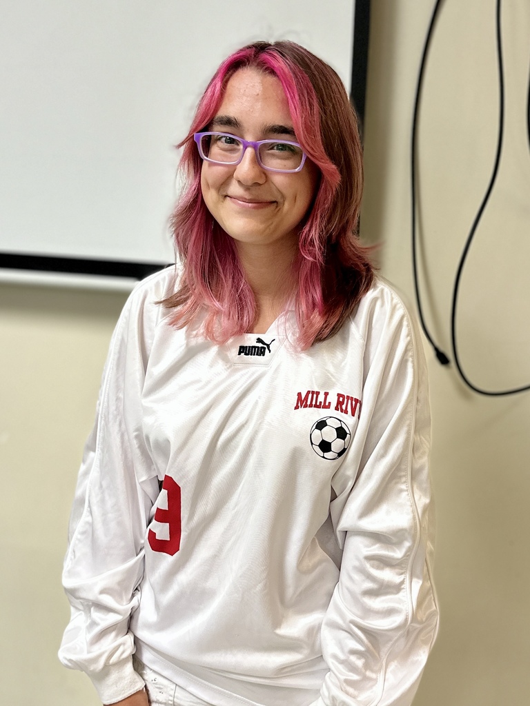 A student with pink hair and glasses smiles while wearing a white Mill River soccer jersey with a Puma logo and soccer ball graphic.