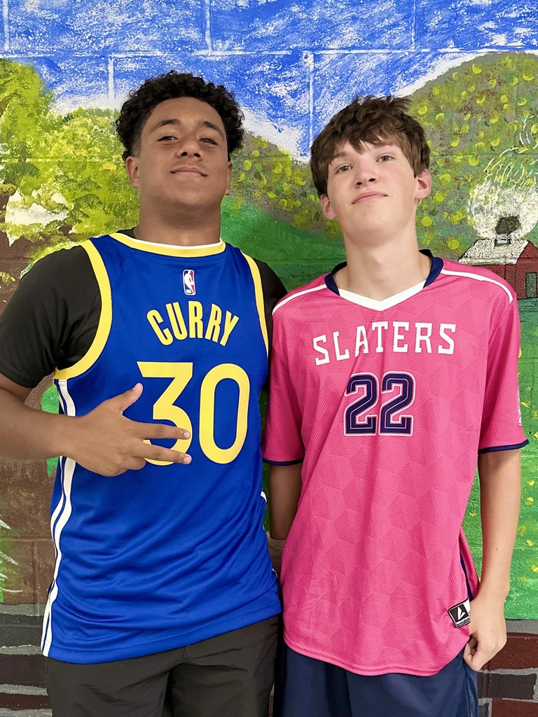 Two students stand side by side, one wearing a blue Golden State Warriors Curry basketball jersey and the other wearing a pink “Slaters” jersey with the number 22, smiling against a mural backdrop.