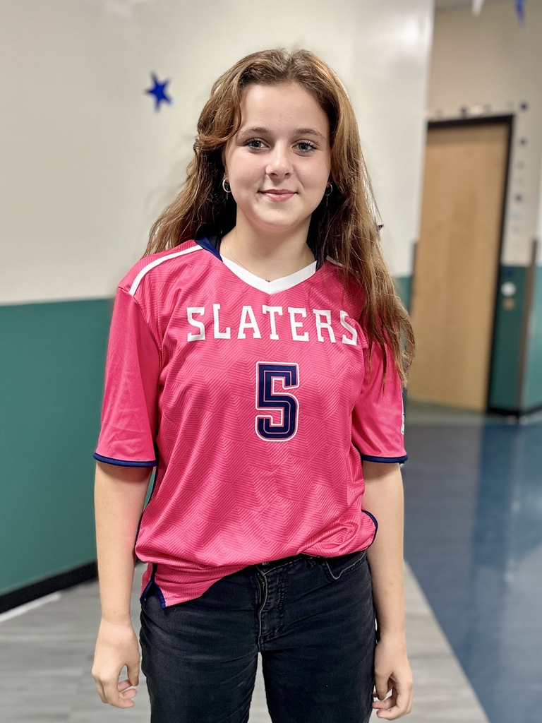 A student stands in a hallway wearing a bright pink “Slaters” jersey with the number 5, smiling at the camera.