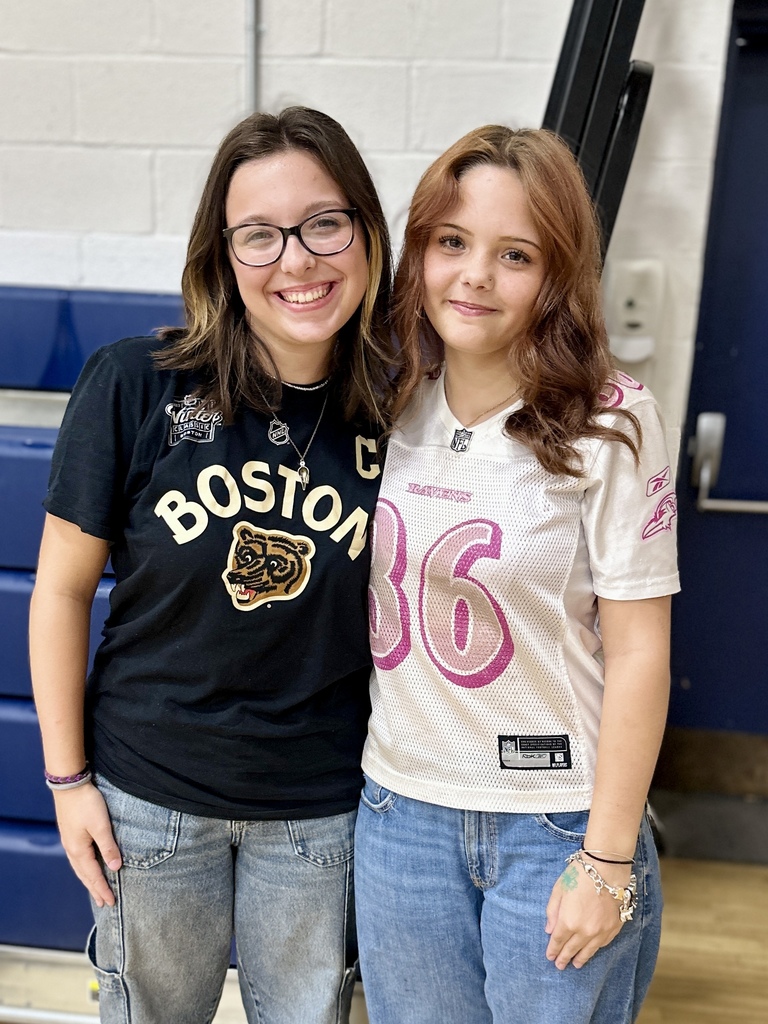 Two students smile and stand arm in arm, one wearing a black Boston Bruins shirt and the other in a white football jersey with pink numbers.