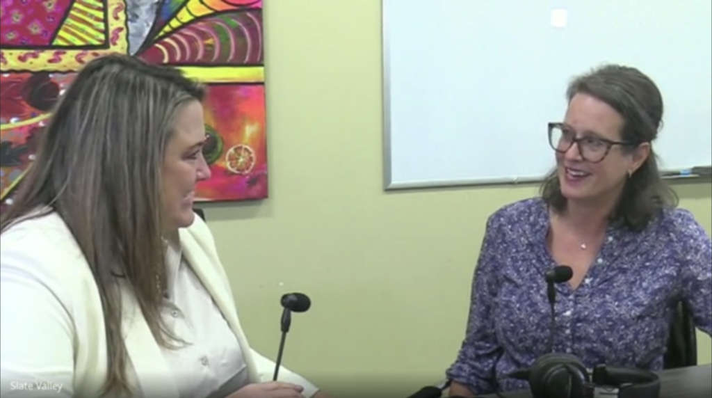 Two women sit across from each other in conversation, both smiling. One wears a white blazer and the other a purple patterned blouse with glasses. They are seated at a table with microphones and a headset in front of them, with a colorful abstract painting and a whiteboard in the background.