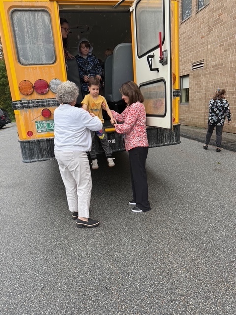 A child in a yellow sweatshirt is helped off the school bus by two adults as classmates wait inside.