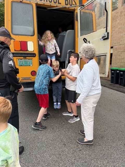Children exit the back of a yellow school bus during a safety drill, guided by an adult with curly gray hair and a police officer standing nearby.