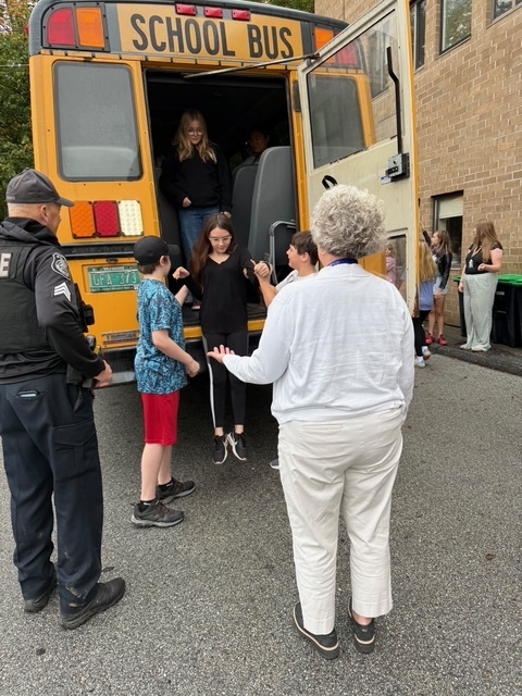 Children leave the bus one by one, guided by adults standing behind and beside the vehicle.