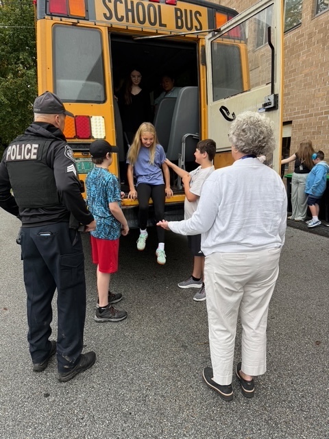 Students practice exiting a school bus during a drill, with adults and a police officer helping them down.