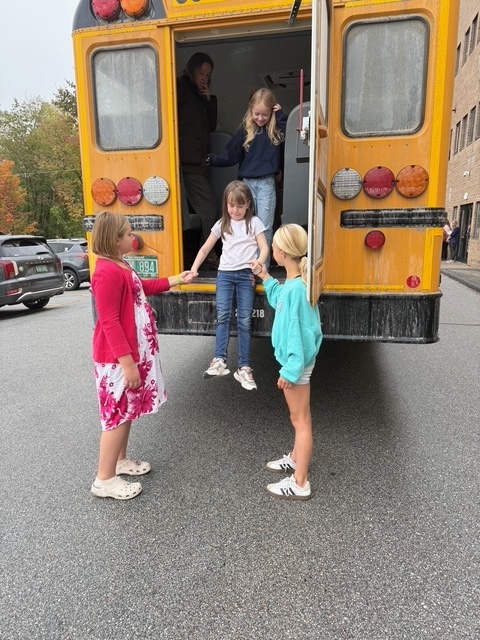 Young students climb out of the back of a school bus, assisted by two older students, one wearing a pink floral dress.