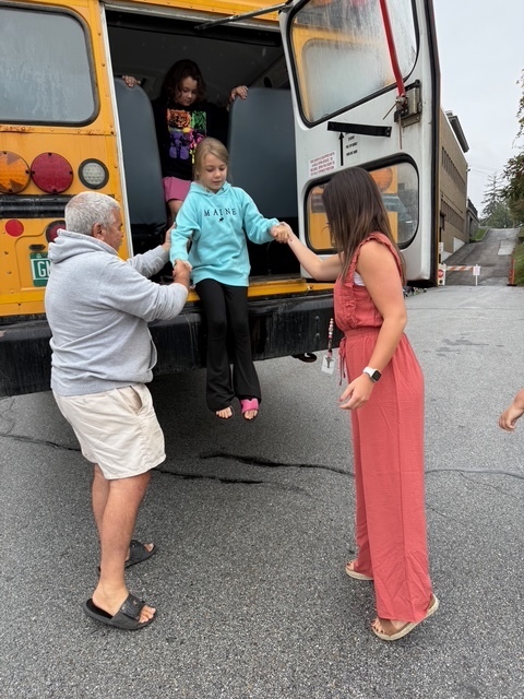 A child wearing a blue hoodie is helped off the back of a bus by two adults, one in a coral jumpsuit.