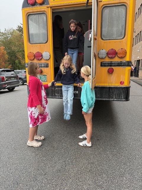 Children step down from a school bus rear exit while two older students hold their hands for safety.