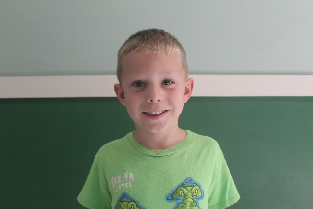  Portrait of a young boy with short, light brown hair, wearing a bright lime green t-shirt with a graphic. He is smiling.