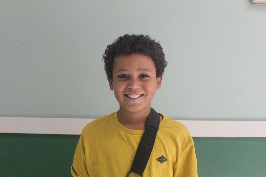 Portrait of a young boy with short, dark, curly hair, wearing a bright yellow t-shirt and a black strap across his shoulder. He is smiling widely.