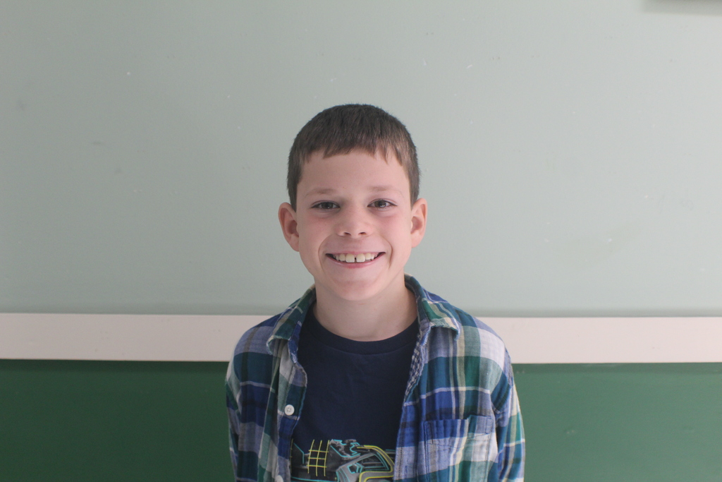 Portrait of a young boy with short, dark hair, wearing a plaid button-up shirt over a dark t-shirt. He is smiling.