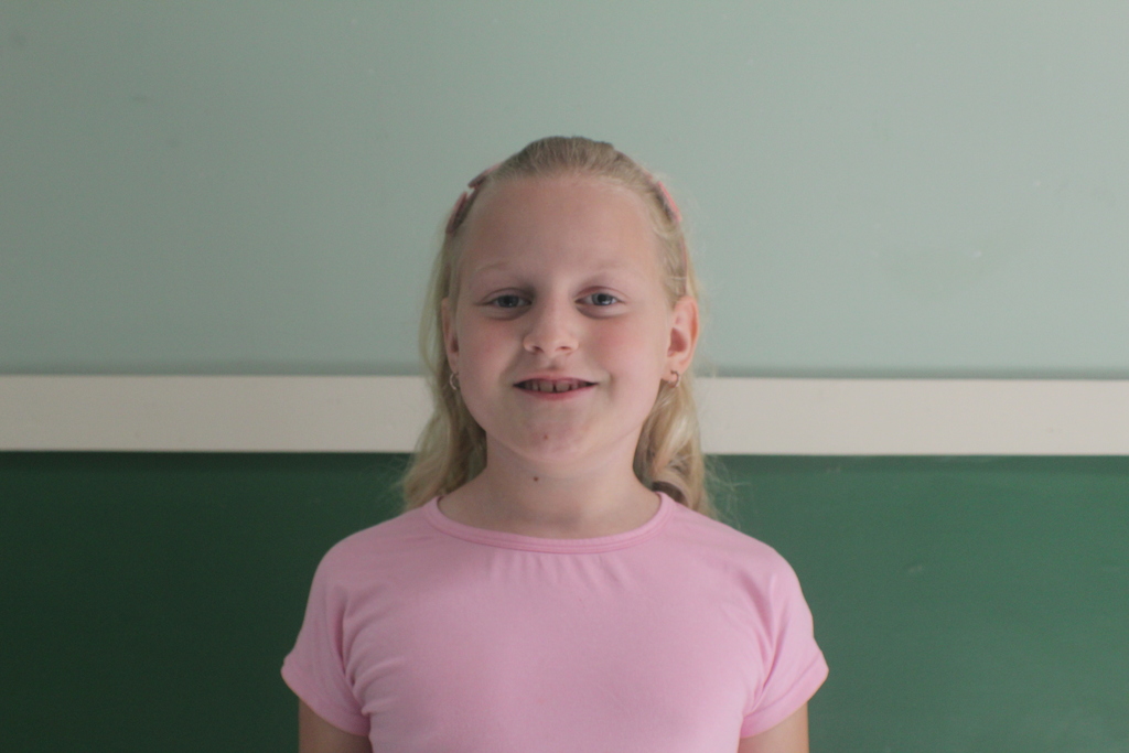 Portrait of a young girl with long blonde hair and a pink headband, wearing a plain pink t-shirt. She is smiling.