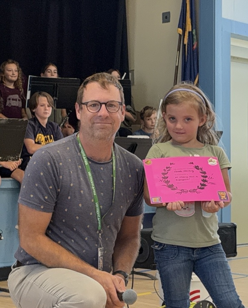 A young female student holding a pink certificate, standing next to a kneeling male staff member wearing a patterned blue t-shirt and glasses. A microphone is in the staff member's hand.