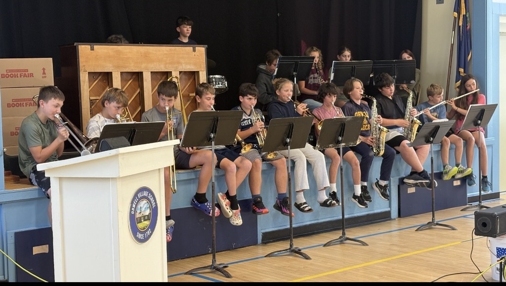 A wide shot of the school band  seated on a stage, playing various instruments including trombones, saxophones, and flutes. The school's sign is visible on the podium. (This is likely the performance component of the assembly.)