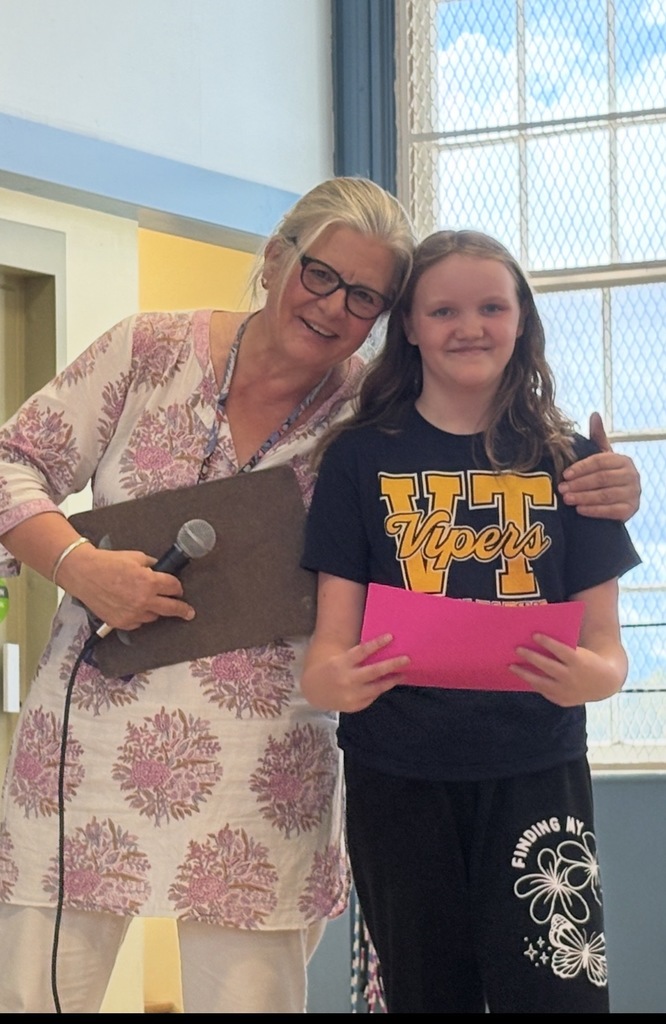 A female student in a dark Vipers t-shirt holds a pink paper, standing next to an older female staff member wearing a white and pink patterned top. The staff member has her arm around the student's shoulder.