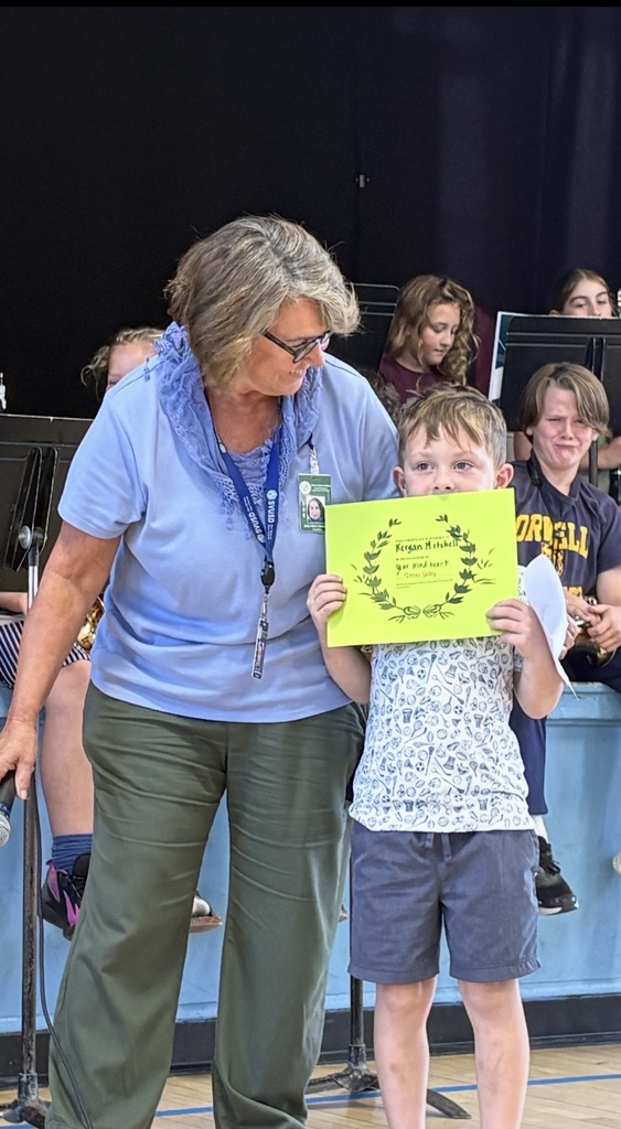 A young male student holding a bright green certificate, looking up at an older female staff member wearing a light blue top and glasses. They are on a stage with other students visible.