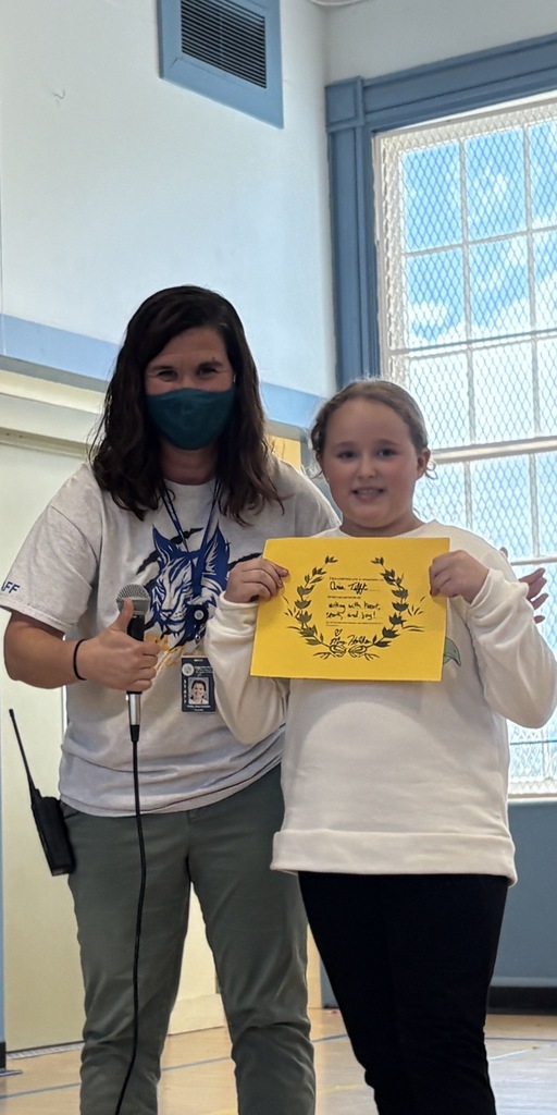 A female student holding a yellow certificate, smiling next to a female staff member wearing a mask and a light-colored t-shirt with a blue mascot graphic. A window is visible behind them.
