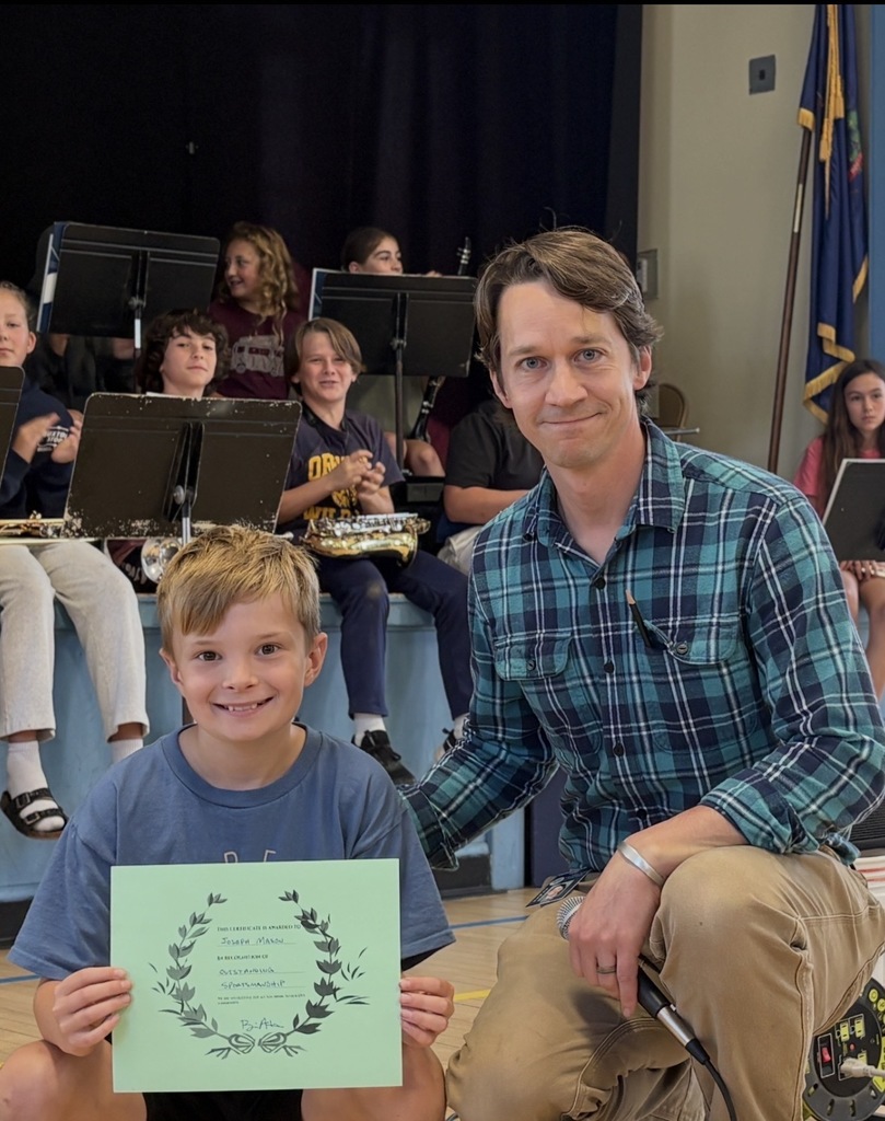 A young male student holding a green certificate with a wreath design, smiling next to a kneeling male staff member wearing a plaid shirt. Other students and music stands are visible in the background.
