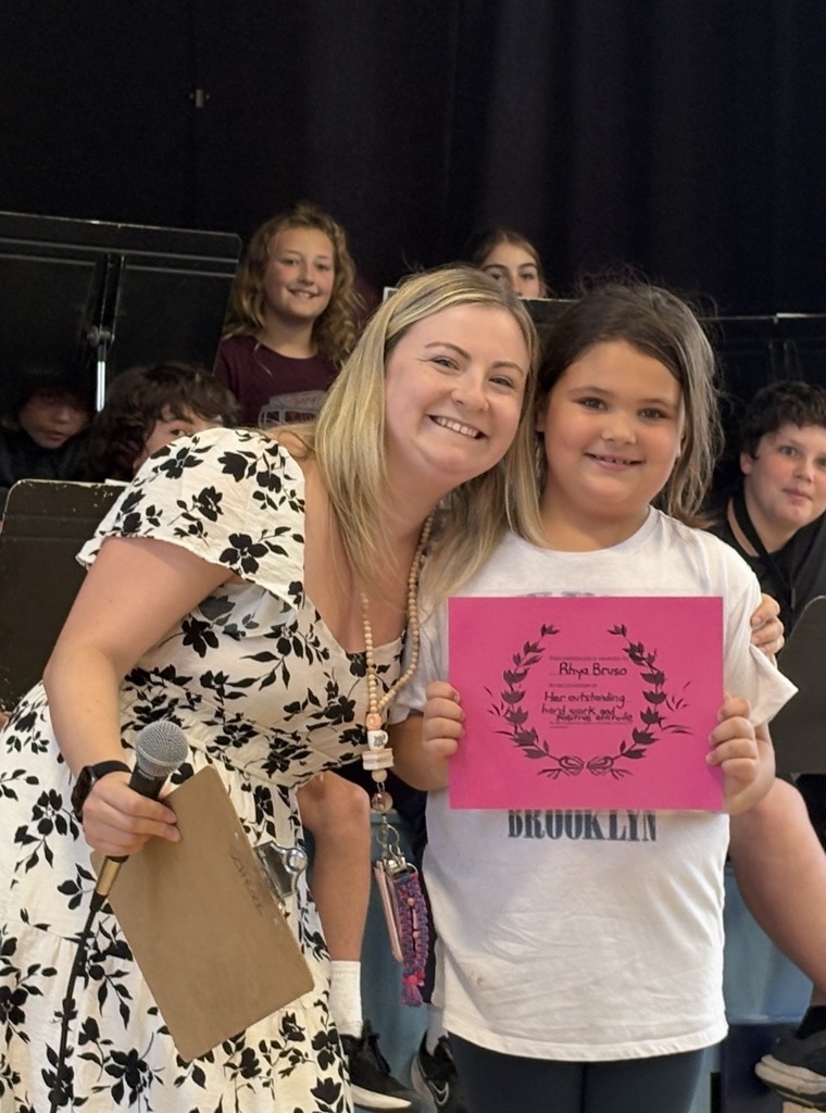 A young female student holding a pink certificate with script, smiling next to a female staff member wearing a white and black floral dress. They are standing on a stage with other students visible behind them.