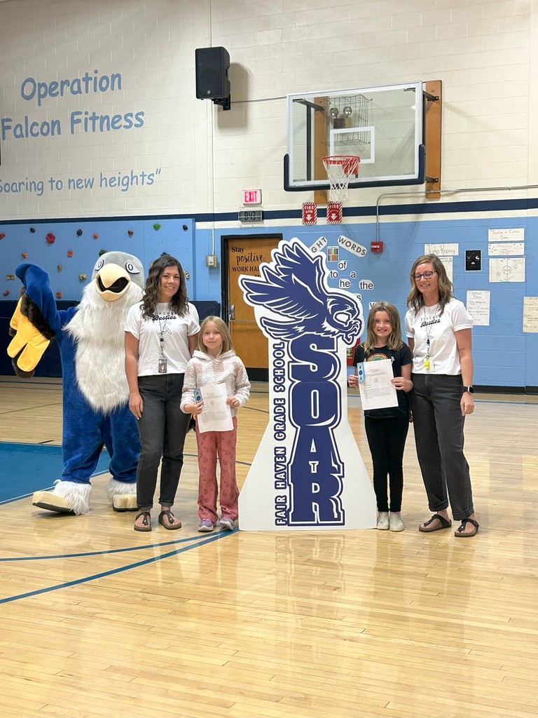 Two students gr. 3 holding certificates stand with two teachers, a falcon mascot, and a large SOAR sign in a school gymnasium.
