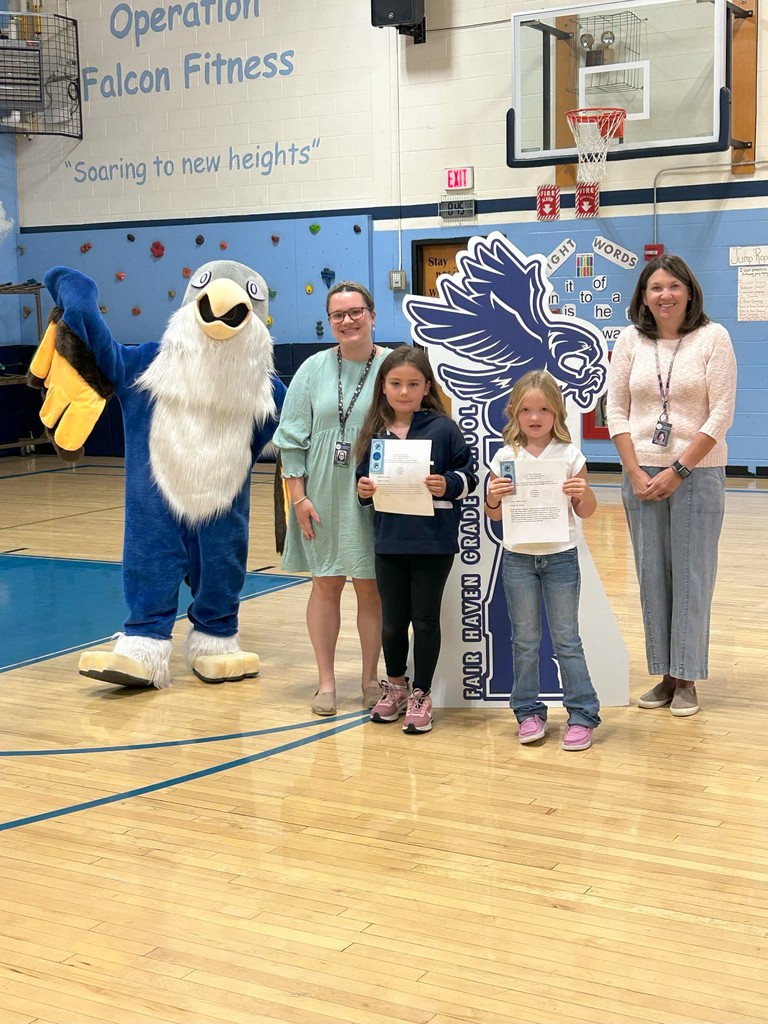 Two students grade 2 holding certificates stand with two teachers, a falcon mascot, and a large SOAR sign in a school gymnasium.