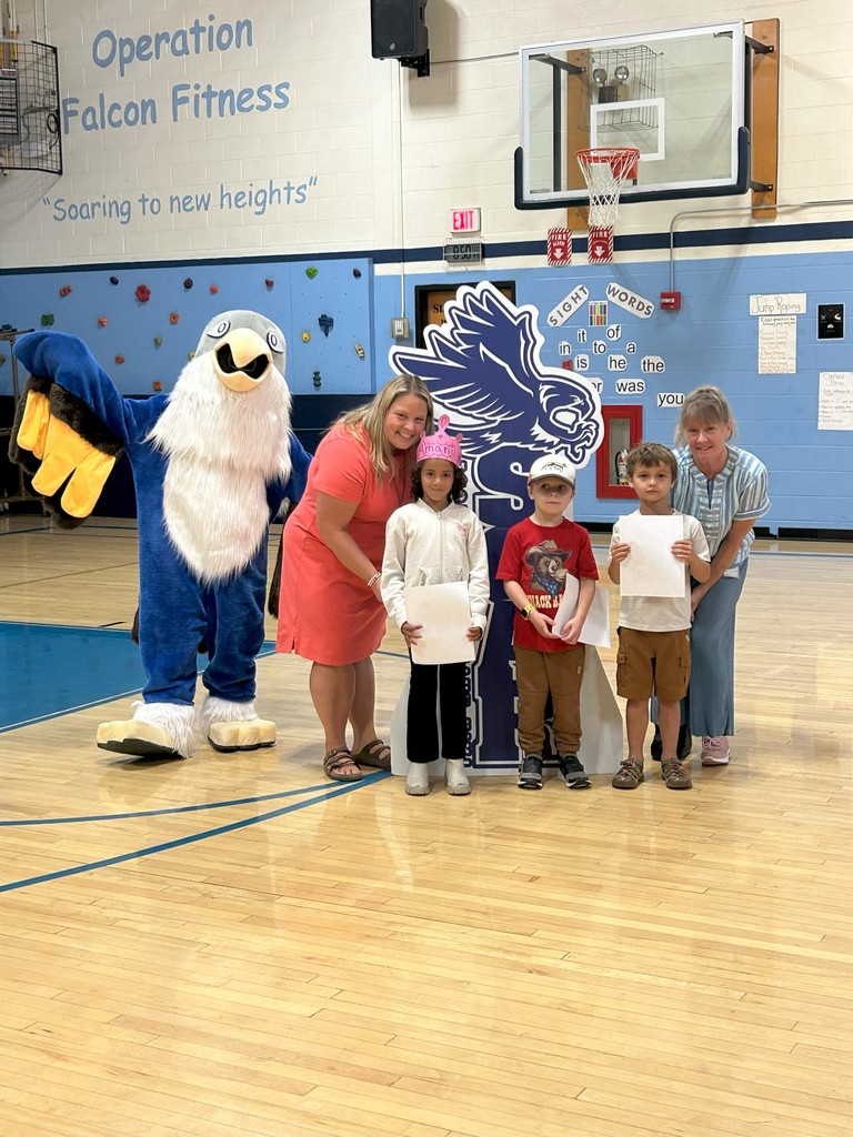 Two students gr 1 holding certificates stand with two teachers, a falcon mascot, and a large SOAR sign in a school gymnasium.