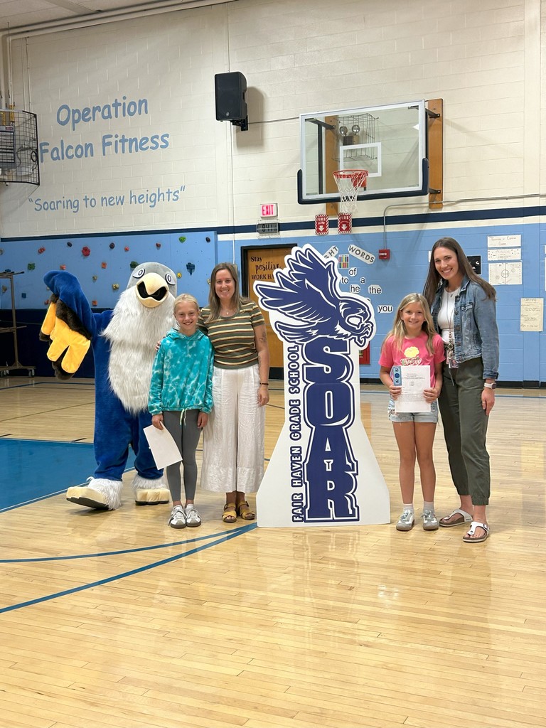 Two students gr 4 holding certificates stand with two teachers, a falcon mascot, and a large SOAR sign in a school gymnasium.