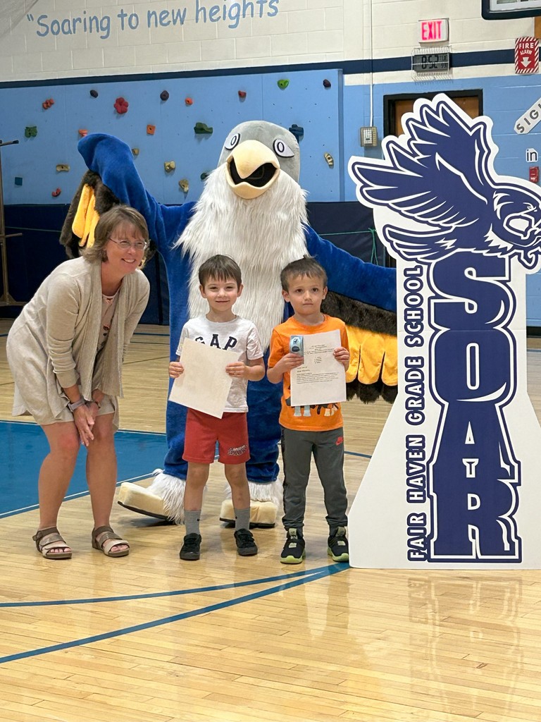 Two students kindergarteners holding certificates stand with two teachers, a falcon mascot, and a large SOAR sign in a school gymnasium.
