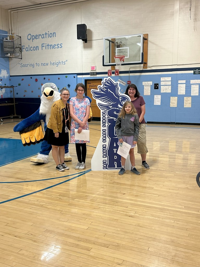 Two students gr. 6 holding certificates stand with two teachers, a falcon mascot, and a large SOAR sign in a school gymnasium.