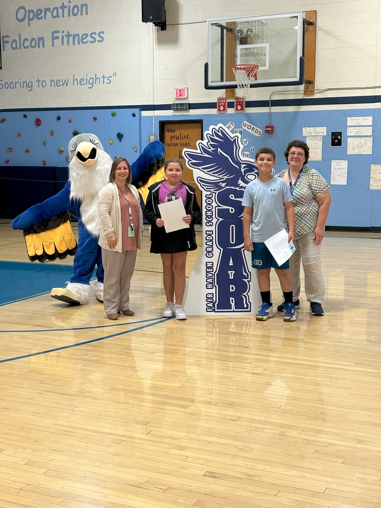 Two students gr 5 holding certificates stand with two teachers, a falcon mascot, and a large SOAR sign in a school gymnasium.
