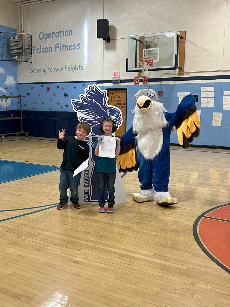 Two health class students holding certificates stand with a falcon mascot, and a large SOAR sign in a school gymnasium.
