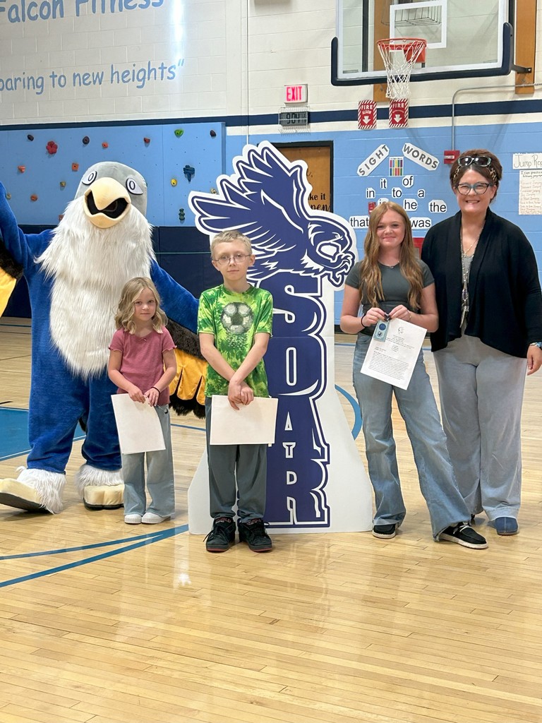 Three students holding certificates stand with art teacher, a falcon mascot, and a large SOAR sign in a school gymnasium.