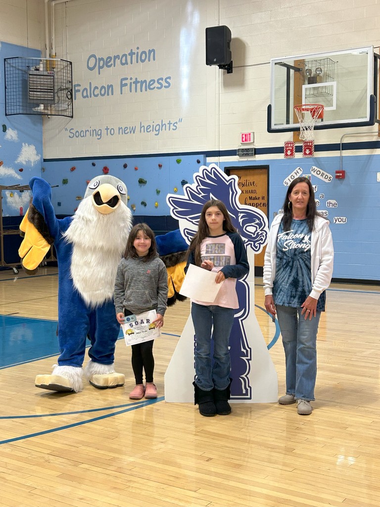 Two students holding bus award certificates stand with a teacher, a falcon mascot, and a large SOAR sign in a school gymnasium.