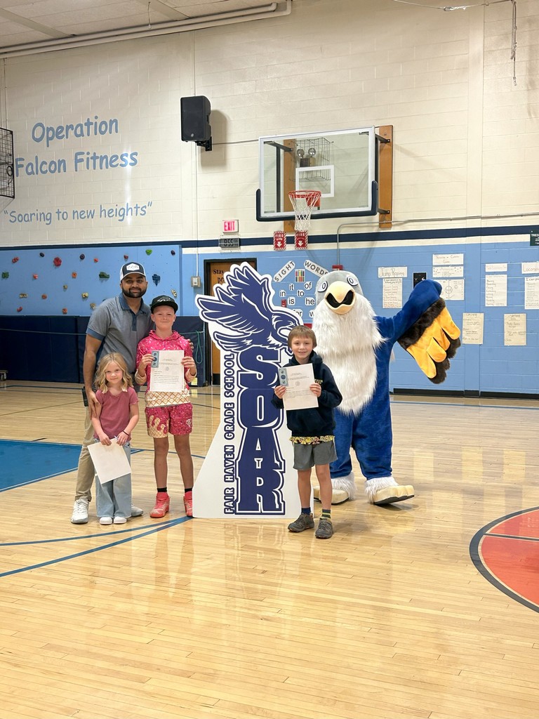 Three students holding certificates stand with the physical education teacher, a falcon mascot, and a large SOAR sign in a school gymnasium.