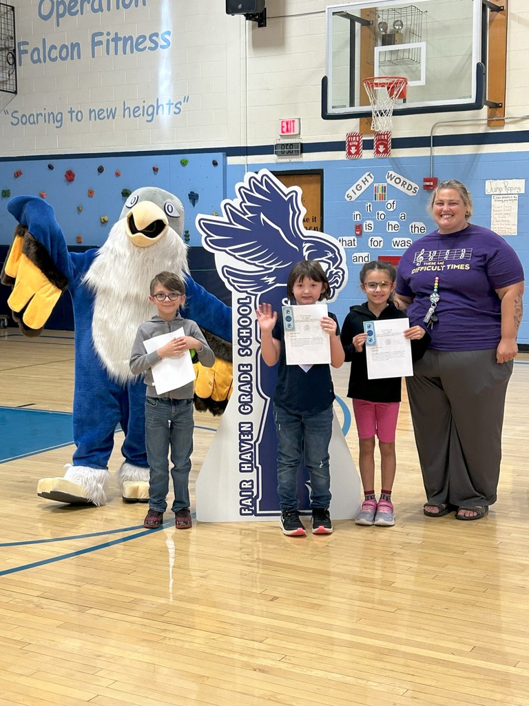 Three students holding certificates stand with the music teacher, a falcon mascot, and a large SOAR sign in a school gymnasium.