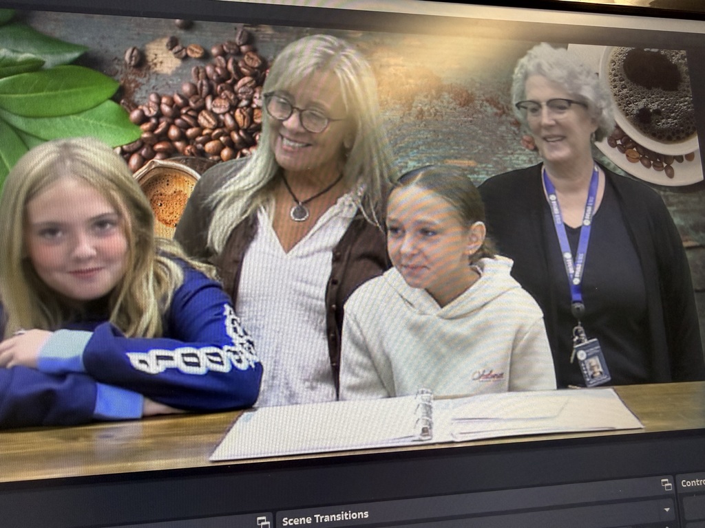 A group of people, including two young girls and two adult women, are sitting at a table in front of a green screen with a coffee-themed background. One of the girls and one of the women are looking at the camera and smiling.