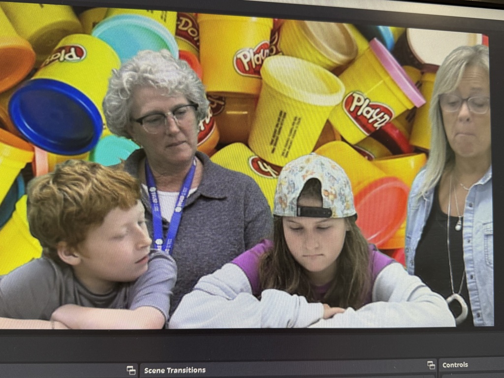 A computer screen displays two students sitting at a table with two adult women. The background shows an array of colorful Play-Doh containers.