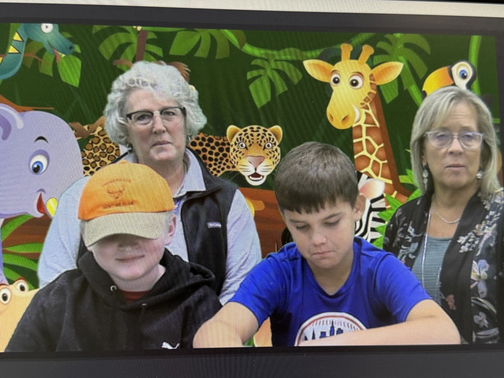 A computer screen shows two boys and two women in front of a green screen background with illustrations of jungle animals.