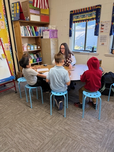A teacher sits at a round table with four young students in a classroom, engaged in a small group activity.